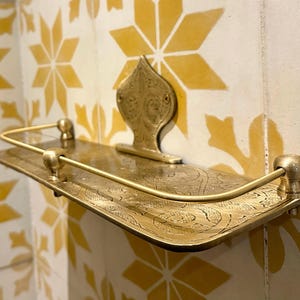 Puede incluir: Estante de pared de color lat&oacute;n con un dise&ntilde;o ornamentado. El estante tiene una base rectangular con grabado decorativo y una barandilla curva. Una placa trasera decorativa con un dise&ntilde;o floral sostiene el estante. El estante est&aacute; montado en una pared alicatada con baldosas amarillas y blancas en forma de estrella.
