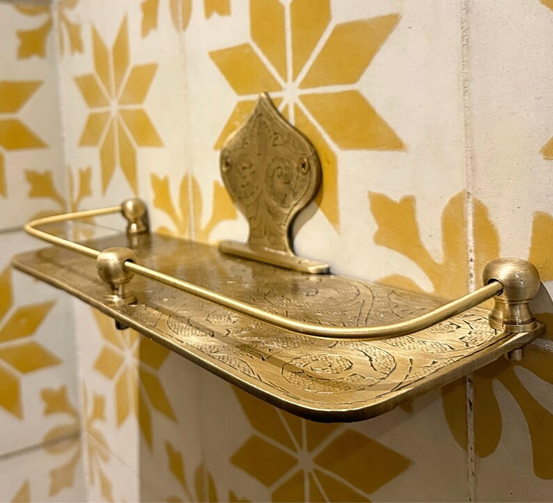 Puede incluir: Estante de pared de color lat&oacute;n con un dise&ntilde;o ornamentado. El estante tiene una base rectangular con grabado decorativo y una barandilla curva. Una placa trasera decorativa con un dise&ntilde;o floral sostiene el estante. El estante est&aacute; montado en una pared alicatada con baldosas amarillas y blancas en forma de estrella.