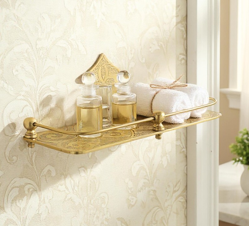 Puede incluir: Estante de ba&ntilde;o de metal dorado con detalles ornamentados. El estante contiene dos botellas de vidrio con tapones transparentes, un vaso peque&ntilde;o y dos toallas blancas enrolladas atadas con hilo. El estante est&aacute; montado en una pared con papel pintado floral.