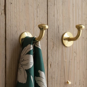 Könnte beinhalten: Zwei Messing-Wandhaken an einer Holzoberfläche. Ein Haken hält einen grünen Schal mit weißem Blumenmuster. Eine goldene Halskette hängt am anderen Haken. Die Haken haben ein gebogenes Design.
