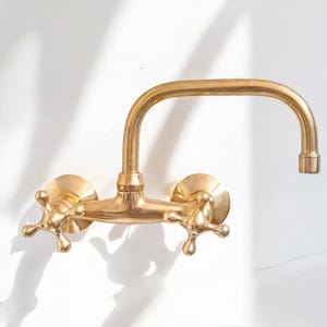 May include: A gold-toned wall-mounted faucet with two cross-shaped handles and a curved spout.