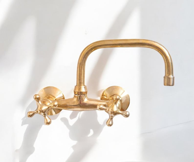 May include: A gold-toned wall-mounted faucet with two cross-shaped handles and a curved spout.