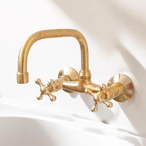 May include: A gold-toned wall-mounted faucet with two cross-shaped handles. The faucet has a curved spout and is designed for a bathroom or kitchen sink.