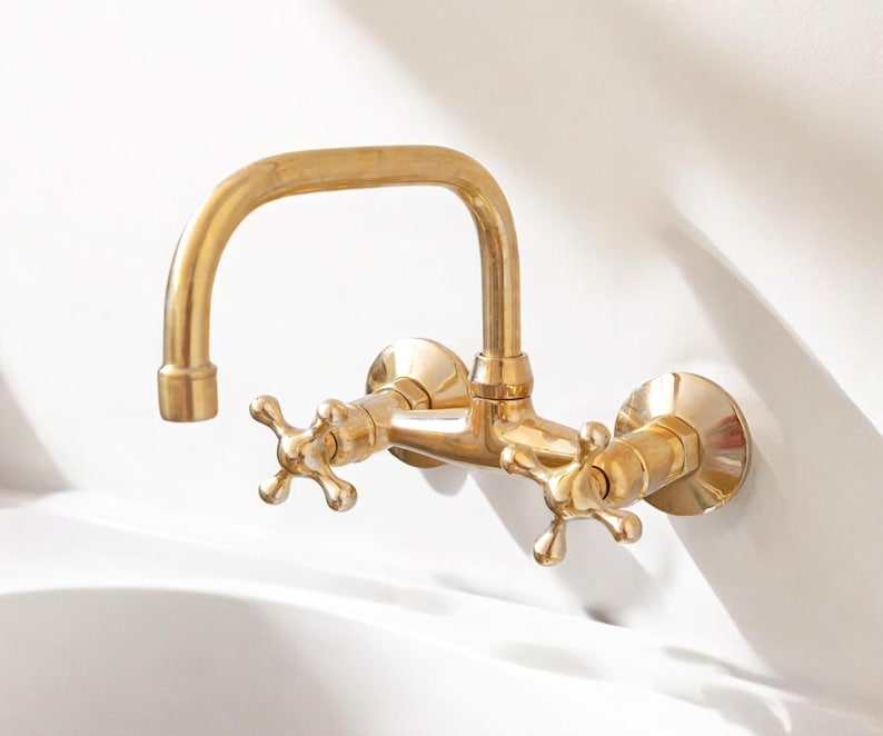 May include: A gold-toned wall-mounted faucet with two cross-shaped handles. The faucet has a curved spout and is designed for a bathroom or kitchen sink.