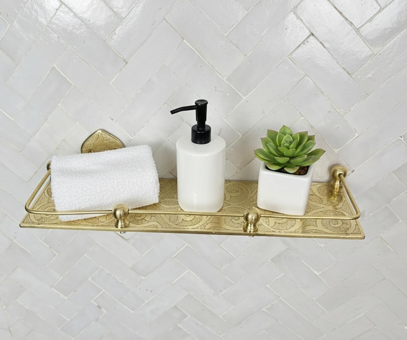 Puede incluir: Un estante de metal dorado con un patr&oacute;n decorativo sostiene una toalla de mano blanca, un dispensador de jab&oacute;n blanco con una bomba negra y una peque&ntilde;a suculenta en maceta. El estante est&aacute; montado en una pared de azulejos blancos.