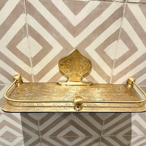 Puede incluir: Estante de metal dorado con intrincados dise&ntilde;os florales. El estante tiene una placa trasera decorativa y soportes curvos. Montado en una pared de azulejos estampados, a&ntilde;ade elegancia a cualquier espacio. Aproximadamente 30 cm de ancho.