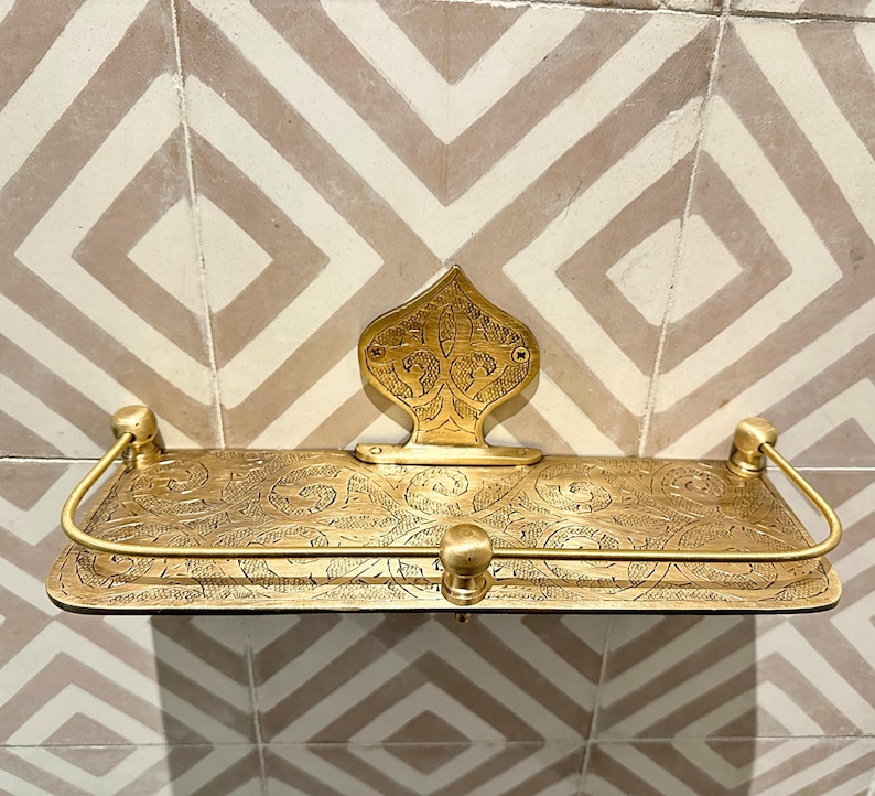 Puede incluir: Estante de metal dorado con intrincados dise&ntilde;os florales. El estante tiene una placa trasera decorativa y soportes curvos. Montado en una pared de azulejos estampados, a&ntilde;ade elegancia a cualquier espacio. Aproximadamente 30 cm de ancho.