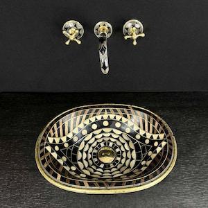K&ouml;nnte beinhalten: Ein ovales Waschbecken in Schwarz und Wei&szlig; mit einem geometrischen Muster. Das Waschbecken hat einen Messingrand und steht auf einer schwarzen Arbeitsplatte. Ein Messinghahn mit schwarzem Griff ist an der Wand &uuml;ber dem Waschbecken montiert.