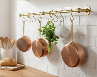 May include: A kitchen scene featuring copper-bottomed pans hanging from a brass rail. A white mug and a bunch of fresh herbs are also suspended. The background is a white tiled wall, and a wooden countertop is in the foreground.