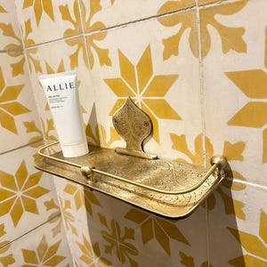Puede incluir: Estante decorativo de lat&oacute;n de estilo antiguo con detalles ornamentados. Sostiene un tubo blanco de protector solar Allie Deluv EX. El estante est&aacute; montado en una pared alicatada con un patr&oacute;n floral amarillo y blanco.