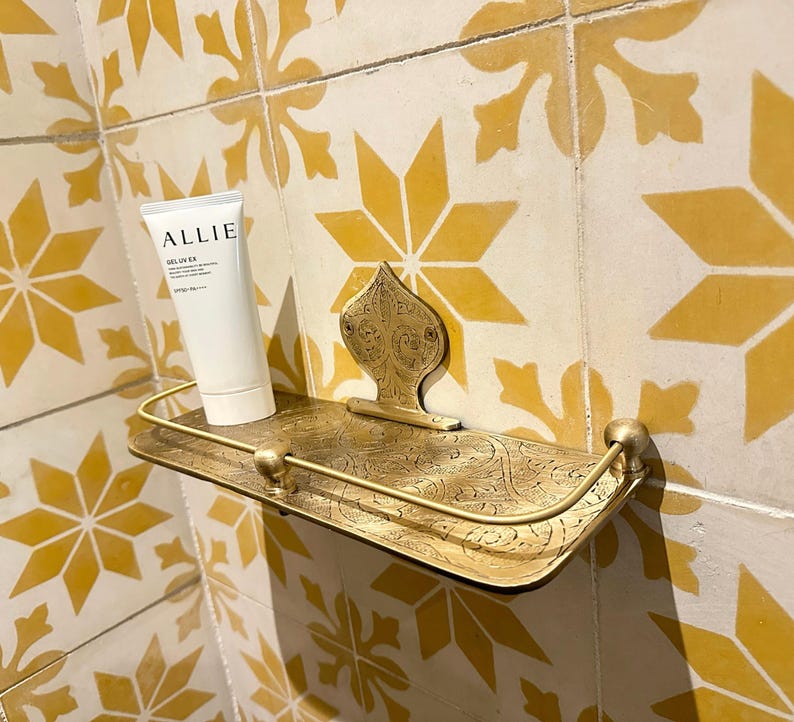 Puede incluir: Estante decorativo de lat&oacute;n de estilo antiguo con detalles ornamentados. Sostiene un tubo blanco de protector solar Allie Deluv EX. El estante est&aacute; montado en una pared alicatada con un patr&oacute;n floral amarillo y blanco.