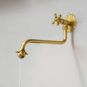 Könnte beinhalten: Ein goldfarbener, an der Wand montierter Wasserhahn mit einer dekorativen floralen Rückplatte. Der Wasserhahn hat einen kreuzförmigen Griff und einen gebogenen Auslauf mit einem kugelförmigen Ende. Wasser fließt aus dem Auslauf.