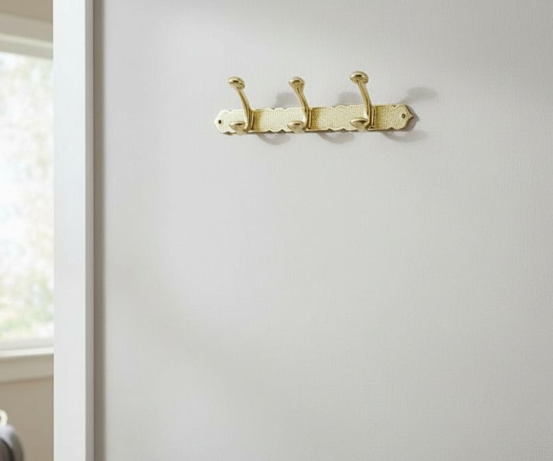 Pode incluir: Um cabide de parede dourado com tr&ecirc;s ganchos. O cabide tem uma borda decorativa, ondulada e &eacute; montado em uma parede branca. Os ganchos s&atilde;o curvos e t&ecirc;m um topo arredondado.