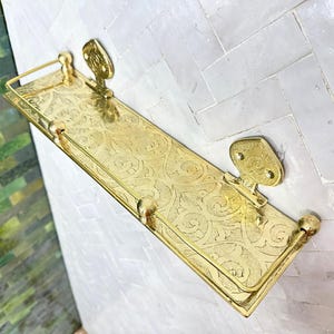 Puede incluir: Una estanter&iacute;a de pared rectangular dorada con un patr&oacute;n ornamental y ondulado. La estanter&iacute;a tiene un borde elevado y est&aacute; soportada por dos soportes decorativos. La estanter&iacute;a es de metal y tiene un acabado pulido. Para ba&ntilde;o o cocina.