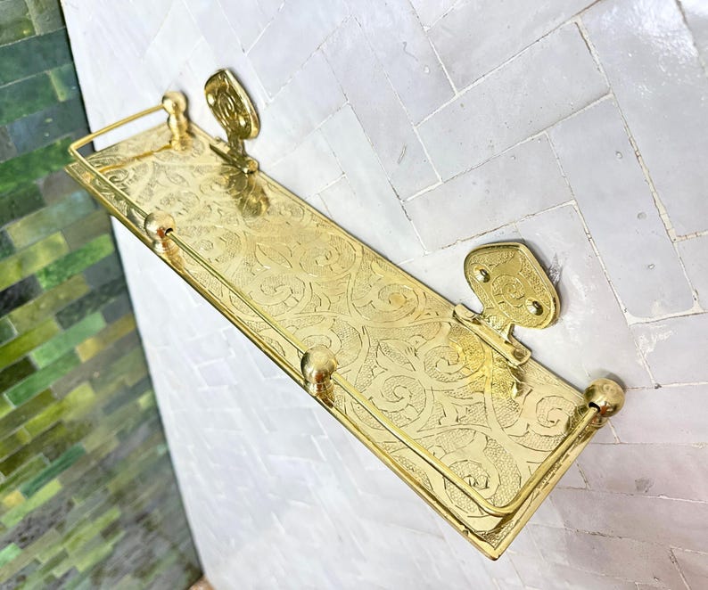 Puede incluir: Una estanter&iacute;a de pared rectangular dorada con un patr&oacute;n ornamental y ondulado. La estanter&iacute;a tiene un borde elevado y est&aacute; soportada por dos soportes decorativos. La estanter&iacute;a es de metal y tiene un acabado pulido. Para ba&ntilde;o o cocina.