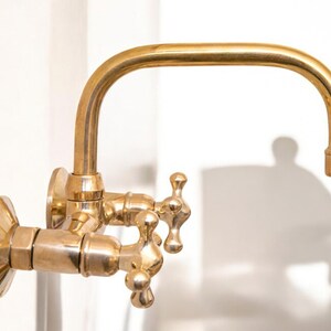 May include: A gold-toned wall-mounted faucet with two handles and a curved spout. The faucet has a traditional design with ornate details.