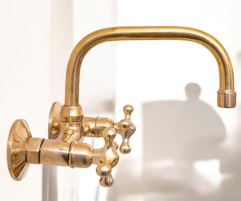 May include: A gold-toned wall-mounted faucet with two handles and a curved spout. The faucet has a traditional design with ornate details.