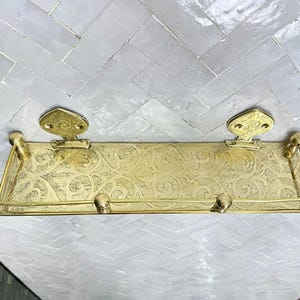 Puede incluir: Un estante de metal dorado con un dise&ntilde;o ornamentado y grabado. El estante tiene dos soportes decorativos y una barra en la parte delantera. El estante est&aacute; montado sobre una superficie de baldosas blancas y grises.