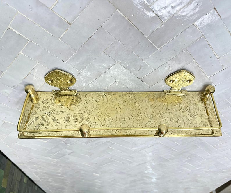 Puede incluir: Un estante de metal dorado con un dise&ntilde;o ornamentado y grabado. El estante tiene dos soportes decorativos y una barra en la parte delantera. El estante est&aacute; montado sobre una superficie de baldosas blancas y grises.
