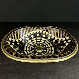 K&ouml;nnte beinhalten: Ovales Waschbecken mit einem geometrischen Muster in Schwarz und Wei&szlig;. Das Waschbecken ist aus Holz und hat einen Messingrand.