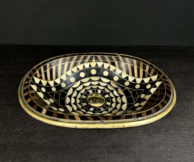 K&ouml;nnte beinhalten: Ovales Waschbecken mit einem geometrischen Muster in Schwarz und Wei&szlig;. Das Waschbecken ist aus Holz und hat einen Messingrand.