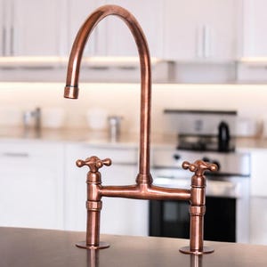 Puede incluir: Un grifo de cocina de cobre con un caño alto y dos manijas de palanca. El grifo está montado en una encimera.