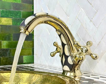 May include: A gold-toned faucet with a black and white patterned design. The faucet has two knobs and is turned on, with water flowing from the spout.