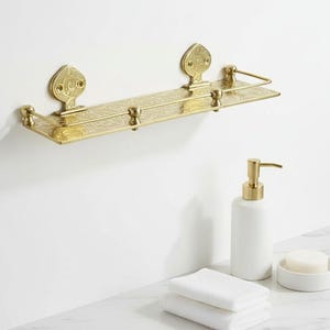 Puede incluir: Estante de ba&ntilde;o dorado y ornamentado con detalles decorativos. El estante est&aacute; montado en una pared blanca. Un dispensador de jab&oacute;n blanco, un jabonera y toallas blancas dobladas est&aacute;n sobre una superficie de m&aacute;rmol.