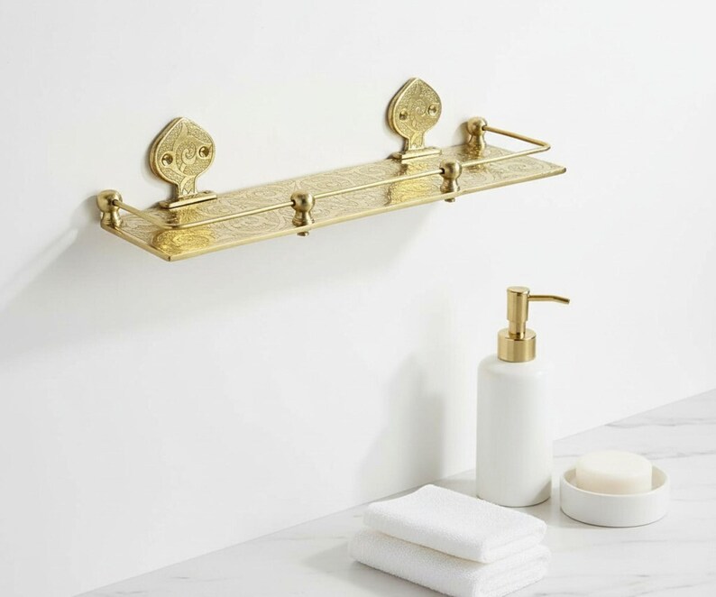Puede incluir: Estante de ba&ntilde;o dorado y ornamentado con detalles decorativos. El estante est&aacute; montado en una pared blanca. Un dispensador de jab&oacute;n blanco, un jabonera y toallas blancas dobladas est&aacute;n sobre una superficie de m&aacute;rmol.