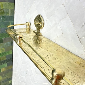 Puede incluir: Una estanter&iacute;a rectangular dorada con intrincados patrones grabados y un acento decorativo en forma de hoja. La estanter&iacute;a tiene una barra en la parte delantera y est&aacute; montada en una pared de azulejos blancos. La estanter&iacute;a mide unos 61 cm de largo.
