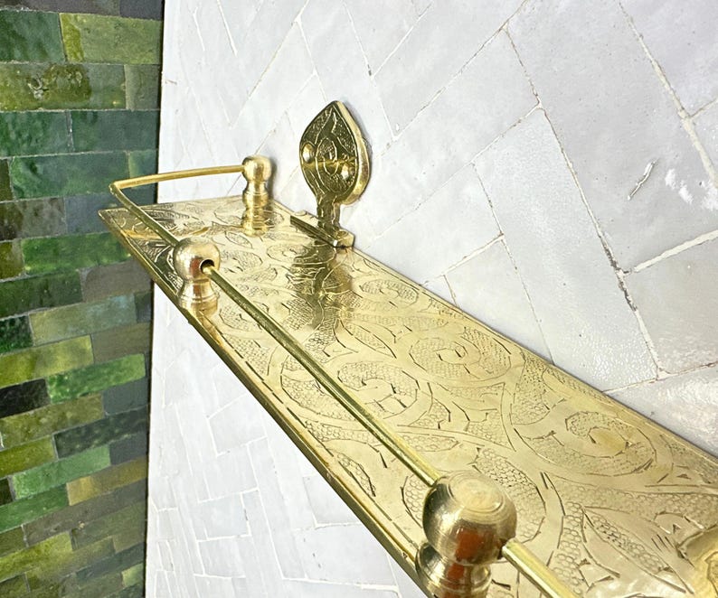 Puede incluir: Una estanter&iacute;a rectangular dorada con intrincados patrones grabados y un acento decorativo en forma de hoja. La estanter&iacute;a tiene una barra en la parte delantera y est&aacute; montada en una pared de azulejos blancos. La estanter&iacute;a mide unos 61 cm de largo.