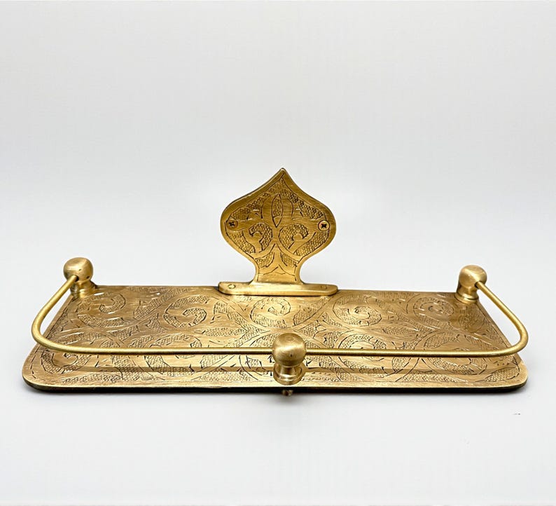 Puede incluir: Un estante rectangular de color lat&oacute;n con un patr&oacute;n floral grabado y ornamentado. El estante tiene una pieza trasera decorativa y dos piezas de extremo redondeadas con una barra curva. Una peque&ntilde;a perilla redonda est&aacute; centrada en la parte delantera.