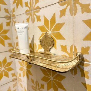 Puede incluir: Un estante de metal dorado decorativo con un dise&ntilde;o ornamentado est&aacute; montado en una pared alicatada. Un tubo blanco de protector solar "ALLIE" est&aacute; en el estante. Los azulejos son amarillos y blancos con un patr&oacute;n de estrella.