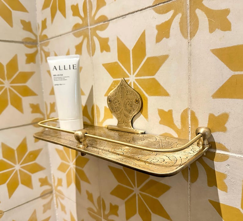 Puede incluir: Un estante de metal dorado decorativo con un dise&ntilde;o ornamentado est&aacute; montado en una pared alicatada. Un tubo blanco de protector solar "ALLIE" est&aacute; en el estante. Los azulejos son amarillos y blancos con un patr&oacute;n de estrella.