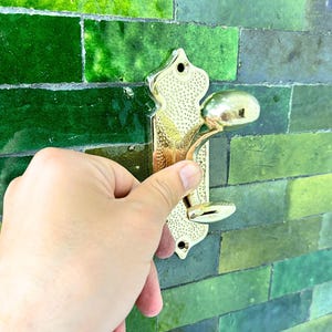 Pode incluir: Um gancho de parede de metal dourado com um design ornamentado. O gancho tem uma placa traseira decorativa e um gancho curvo para pendurar objetos. O fundo &eacute; uma parede de azulejos verdes.
