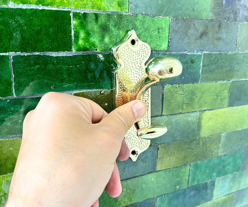 Pode incluir: Um gancho de parede de metal dourado com um design ornamentado. O gancho tem uma placa traseira decorativa e um gancho curvo para pendurar objetos. O fundo &eacute; uma parede de azulejos verdes.