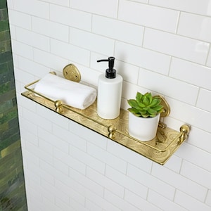 Op de afbeelding: Een goudkleurig metalen badkamerrek met een geëtst ontwerp. Het bevat een witte opgerolde handdoek, een witte zeepdispenser met een zwarte pomp en een kleine vetplant in pot. Het rek is bevestigd aan een wit betegelde muur.
