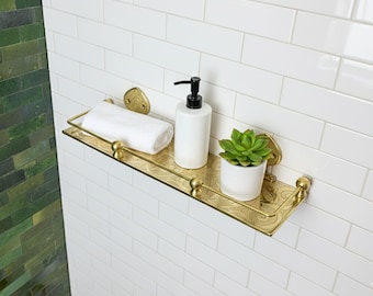May include: A gold-colored metal bathroom shelf with a decorative etched design. The shelf holds a white rolled towel, a white soap dispenser with a black pump, and a small potted succulent. The shelf is mounted on a white tiled wall.