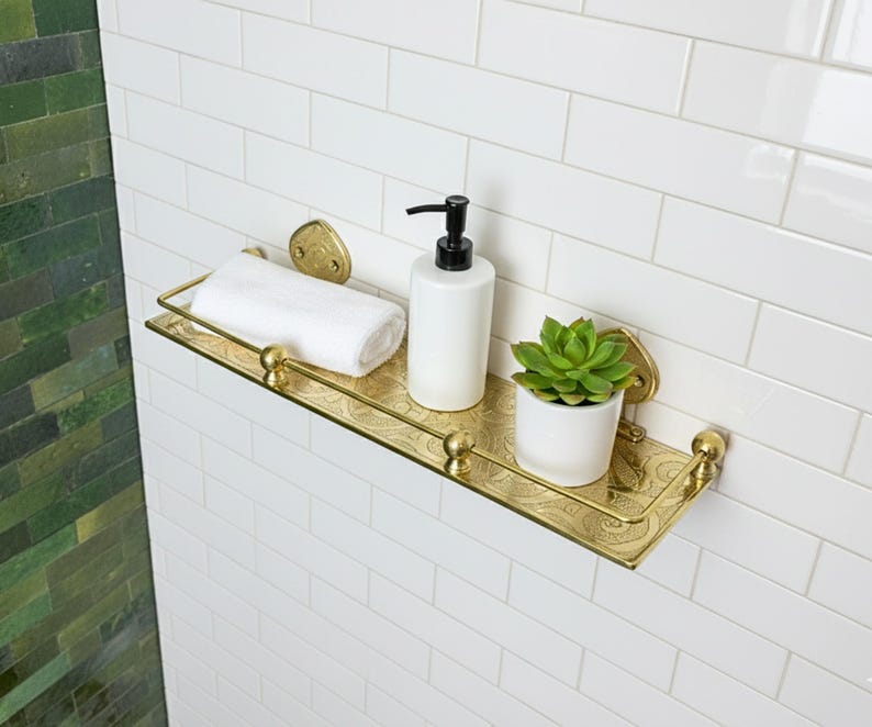 Puede incluir: Estante de ba&ntilde;o de metal dorado con un dise&ntilde;o grabado. Contiene una toalla blanca enrollada, un dispensador de jab&oacute;n blanco con bomba negra y una peque&ntilde;a suculenta en maceta. El estante est&aacute; montado en una pared de azulejos blancos.