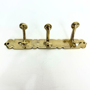 Pode incluir: Um cabide de parede de lat&atilde;o com tr&ecirc;s ganchos curvos. O cabide tem um design decorativo texturizado e uma borda ondulada. Os ganchos s&atilde;o espa&ccedil;ados uniformemente ao longo da barra horizontal, proporcionando uma solu&ccedil;&atilde;o de armazenamento elegante.