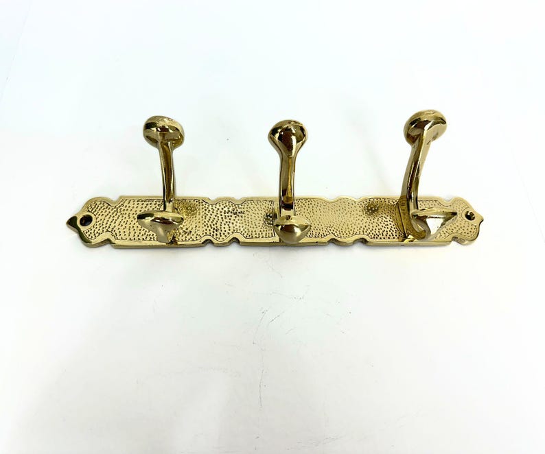 Pode incluir: Um cabide de parede de lat&atilde;o com tr&ecirc;s ganchos curvos. O cabide tem um design decorativo texturizado e uma borda ondulada. Os ganchos s&atilde;o espa&ccedil;ados uniformemente ao longo da barra horizontal, proporcionando uma solu&ccedil;&atilde;o de armazenamento elegante.