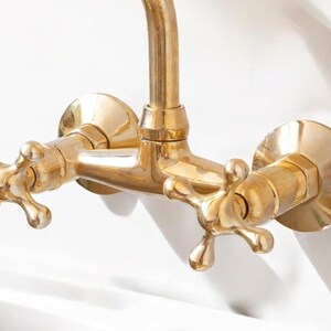 May include: A gold-toned faucet with two cross-shaped handles. The faucet is mounted to a wall and has a spout that extends out.