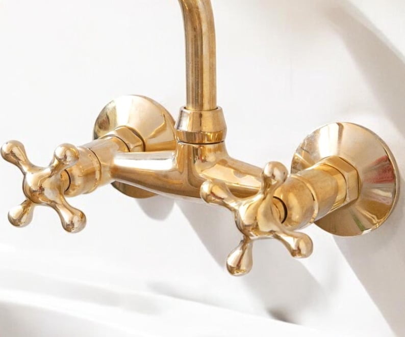 May include: A gold-toned faucet with two cross-shaped handles. The faucet is mounted to a wall and has a spout that extends out.