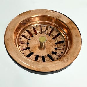 May include: A copper sink strainer with a round, flat design and a central drain hole. The strainer has a series of small holes around the edge and a larger central hole. The strainer is made of copper and has a shiny finish.