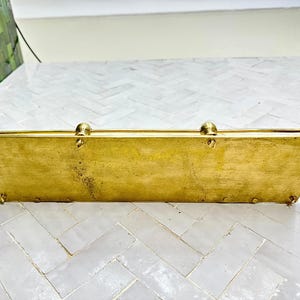 Puede incluir: Una caja rectangular de lat&oacute;n con tapa abisagrada y remates redondeados. La caja tiene una forma ligeramente trapezoidal y peque&ntilde;os pies. El lat&oacute;n tiene un tono dorado c&aacute;lido con algunas zonas de p&aacute;tina, lo que sugiere edad y uso.