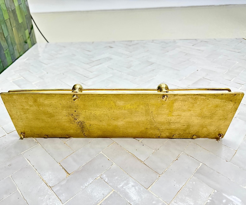 Puede incluir: Una caja rectangular de lat&oacute;n con tapa abisagrada y remates redondeados. La caja tiene una forma ligeramente trapezoidal y peque&ntilde;os pies. El lat&oacute;n tiene un tono dorado c&aacute;lido con algunas zonas de p&aacute;tina, lo que sugiere edad y uso.