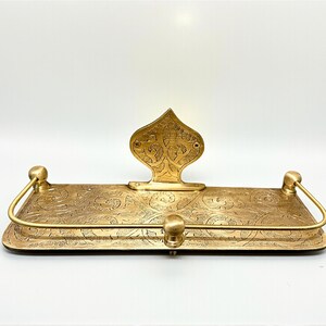 Puede incluir: Un estante de metal dorado con un patr&oacute;n floral grabado y ornamentado. El estante tiene una pieza trasera decorativa y dos asas curvas. El estante est&aacute; dise&ntilde;ado para guardar objetos y a&ntilde;ade un toque de elegancia a cualquier espacio.