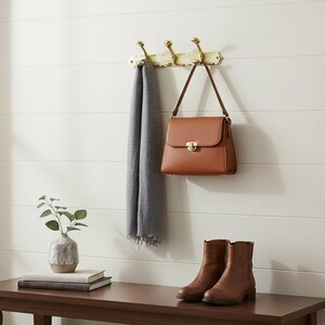 Pode incluir: Uma bolsa de couro castanho com fecho dourado est&aacute; pendurada em um cabide decorativo de lat&atilde;o. Um cachecol cinza est&aacute; pendurado nos ganchos. Abaixo, um par de botas de couro marrom e um pequeno vaso com vegeta&ccedil;&atilde;o est&atilde;o em um banco de madeira.