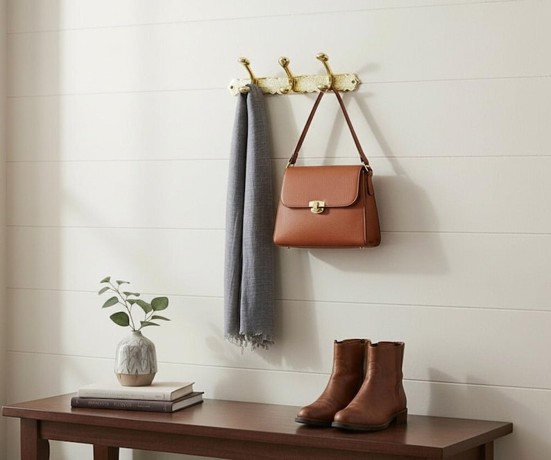 Pode incluir: Uma bolsa de couro castanho com fecho dourado est&aacute; pendurada em um cabide decorativo de lat&atilde;o. Um cachecol cinza est&aacute; pendurado nos ganchos. Abaixo, um par de botas de couro marrom e um pequeno vaso com vegeta&ccedil;&atilde;o est&atilde;o em um banco de madeira.