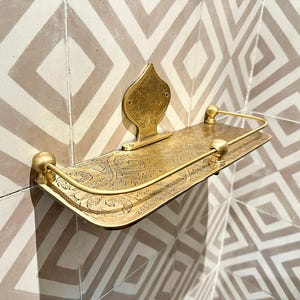 Puede incluir: Un estante dorado montado en la pared con un dise&ntilde;o ornamentado y grabado. El estante tiene un frente curvo con un borde elevado y una placa trasera decorativa. Una barra de metal curvada recorre el borde frontal. El estante est&aacute; montado en una pared de azulejos estampados.